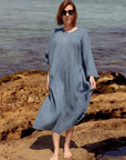 A woman La Bottega di Brunella Linen Dress, long-sleeve blue-gray textured and sunglasses stands barefoot on rocky shoreline with water in the background. A pair of black sandals rests on the rocks beside her.