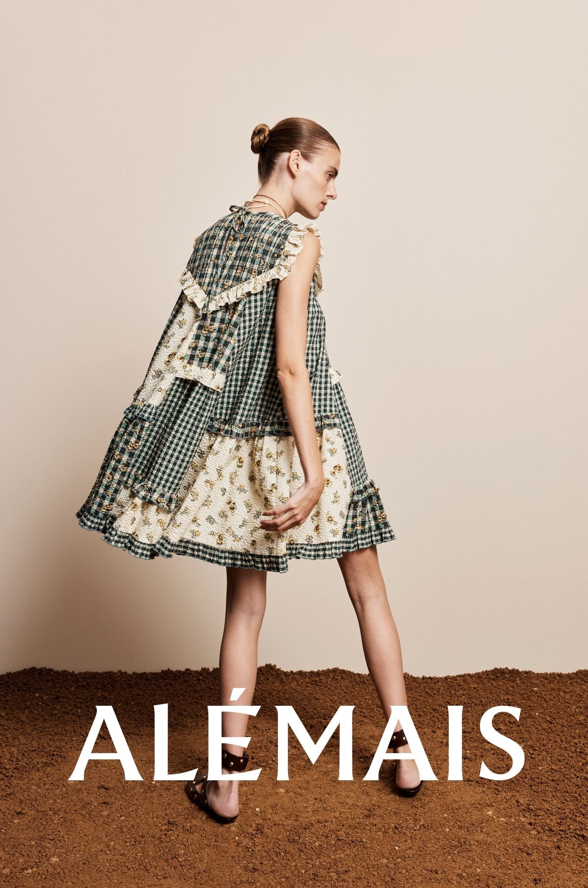 Alemais dress worn by model dress is a sleeveless patchwork dress with a green and cream plaid and floral pattern, set against a beige wall and brown floor. The brand name 'ALEMAIS' is printed in white text in the bottom left corner.