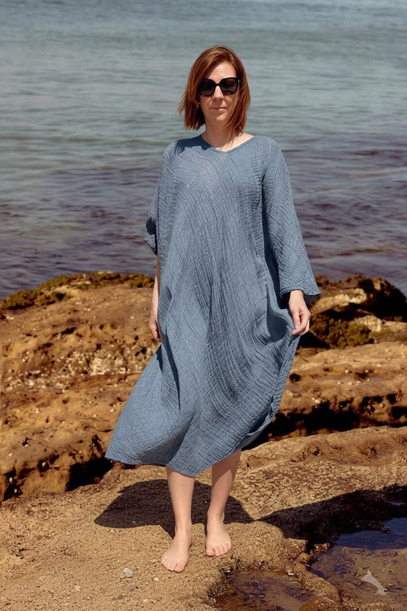 A woman La Bottega di Brunella Linen Dress, long-sleeve blue-gray textured  and sunglasses stands barefoot on rocky shoreline with water in the background. A pair of black sandals rests on the rocks beside her.